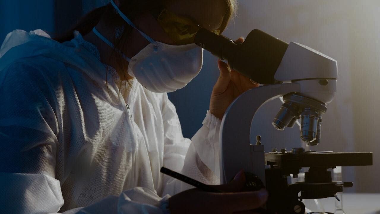 Person wearing a white lab coat and mask, using a microscope in a laboratory setting.